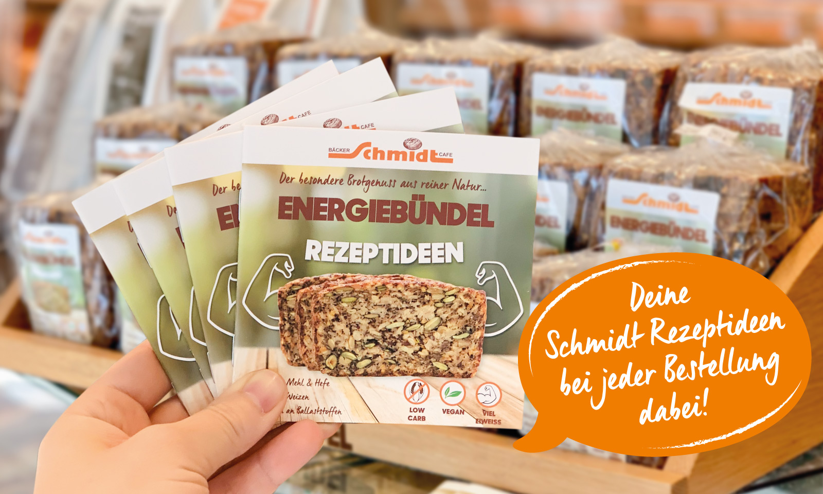 Eine Hand hält 4 kleine Rezeptbücher vom Energiebündel in die Luft. Dahinter befinden sich verpackte Energiebündel auf der Verkaufstheke